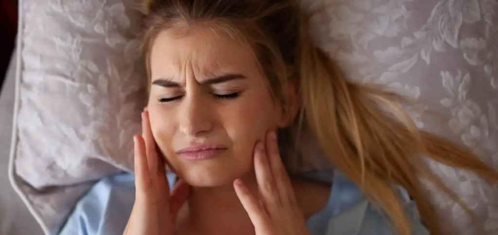 Młoda kobieta z blond włosami leżąca w łóżku na poduszce w kwiatowy wzór, z rękami przy twarzy, wyrażająca ból i dyskomfort, prawdopodobnie spowodowany bruksizmem i zgrzytaniem zębami w nocy. Zdjęcie ilustruje wpływ bruksizmu na zdrowie jamy ustnej przed leczeniem, oferując wsparcie w klinice stomatologicznej SmileClinic