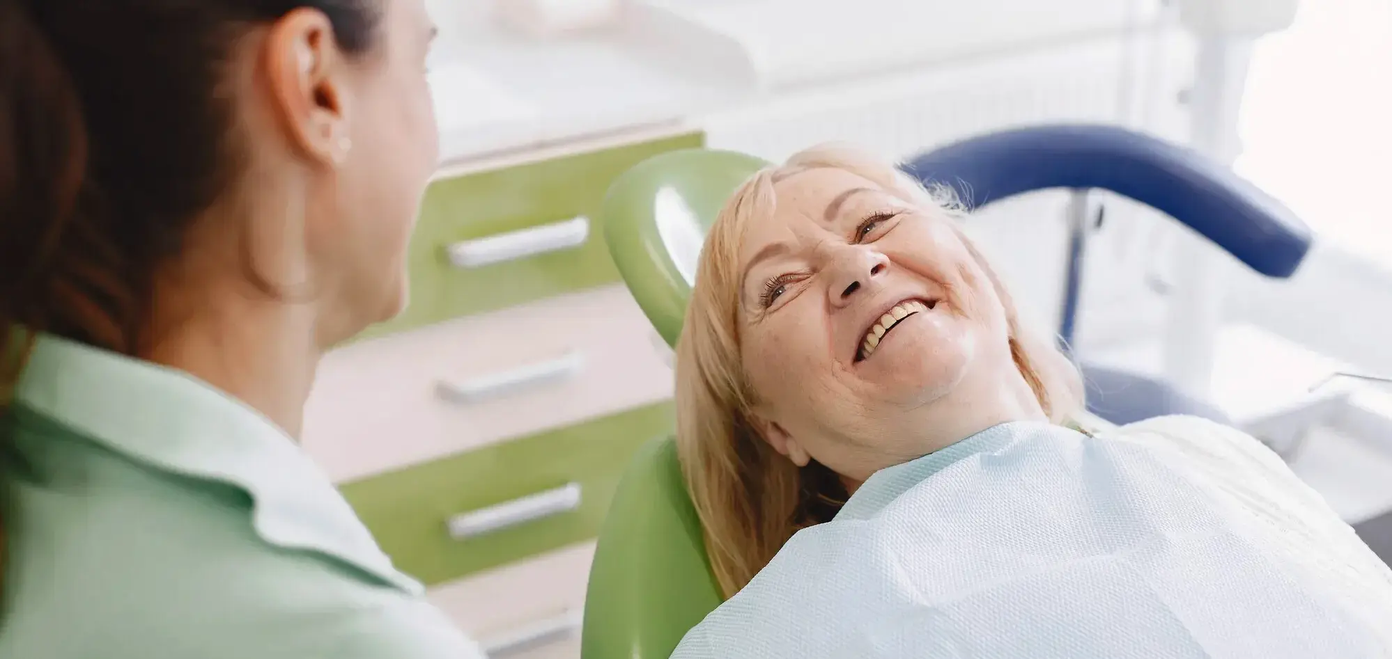Dentystka w zielonej bluzie rozmawia z uśmiechniętą pacjentką leżącą na zielonym fotelu stomatologicznym, w tle widoczne są meble gabinetu. Zdjęcie przedstawia przygotowanie do przebudowy protetycznej, gdzie leczenie endodontyczne jest kluczowym elementem, oferowanym przez klinikę stomatologiczną SmileClinic