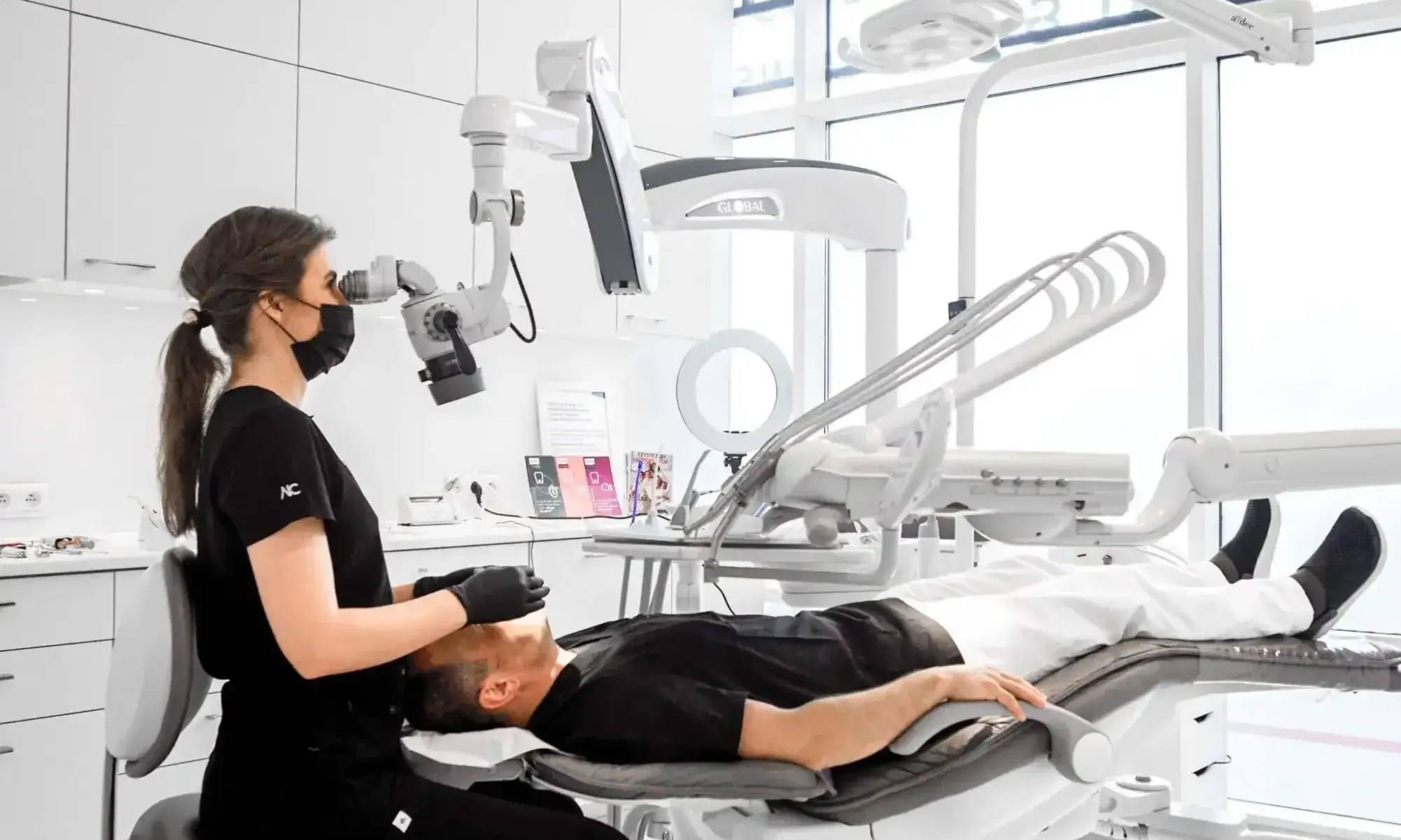 endodontka-pacjentka-przygotowanie-protetyczne-smileclinic-pl kopia Dentystka w czarnej bluzie z logo SmileClinic, z maską i mikroskopem dentystycznym, pracuje przy pacjencie leżącym na fotelu, w nowoczesnym gabinecie z oknami i zaawansowanym sprzętem stomatologicznym. Zdjęcie ilustruje leczenie endodontyczne jako istotny element przygotowania do przebudowy protetycznej, oferowane przez klinikę SmileClinic