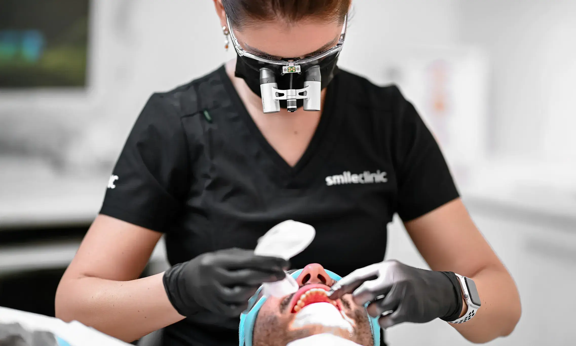 higienistka-leczenie-endodontyczne-przebudowa-protetyczna-smileclinic-pl kopia Higienistka w czarnej bluzie z logo SmileClinic, z okularami powiększającymi i maską, przeprowadza higienizację u pacjenta leżącego na fotelu stomatologicznym, używając narzędzi stomatologicznych. Zdjęcie ilustruje przygotowanie do przebudowy protetycznej, gdzie higienizacja odgrywa ważną rolę w ratowaniu zębów