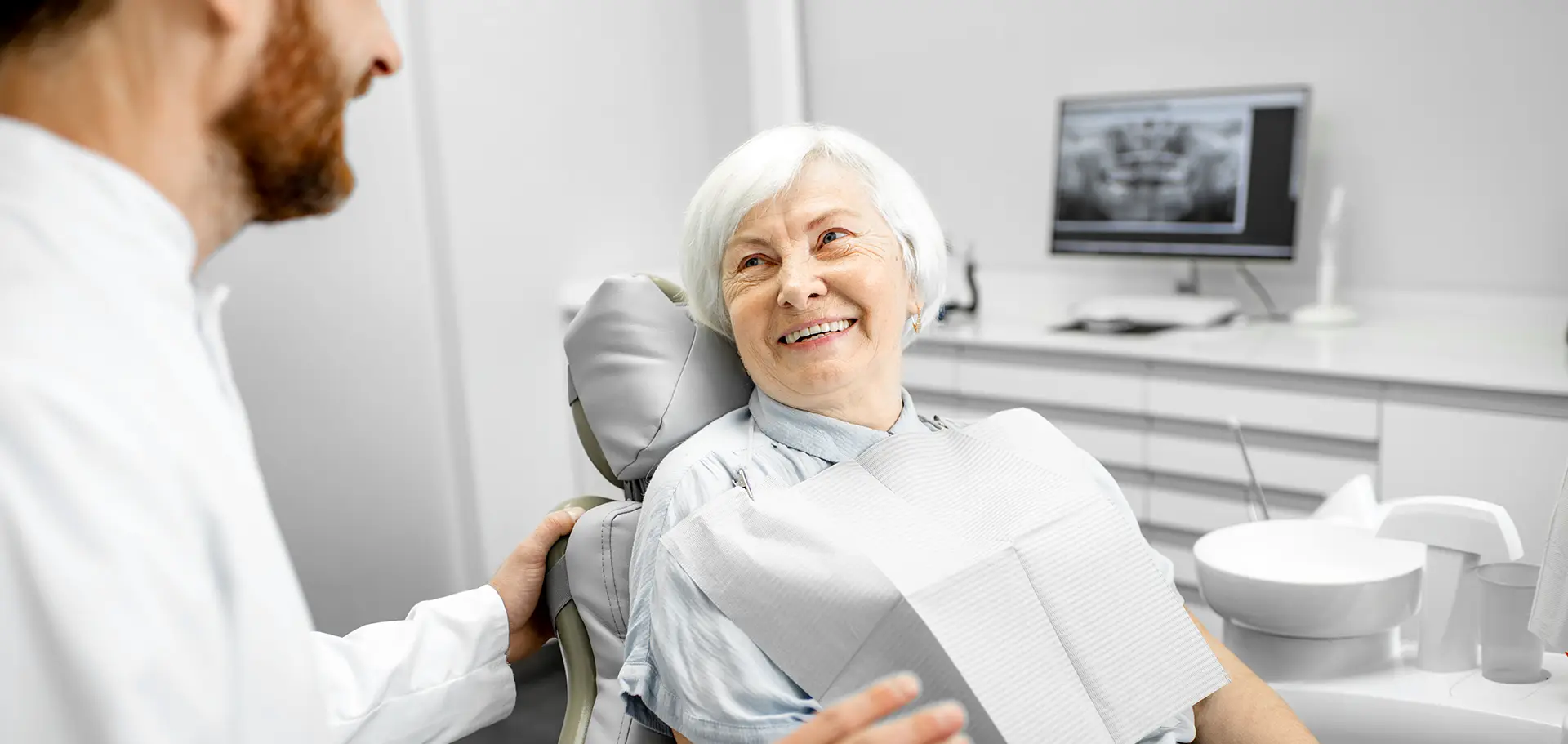 Starsza kobieta z uśmiechem siedzi na fotelu dentystycznym, rozmawiając z lekarzem stomatologiem w nowoczesnym gabinecie.
