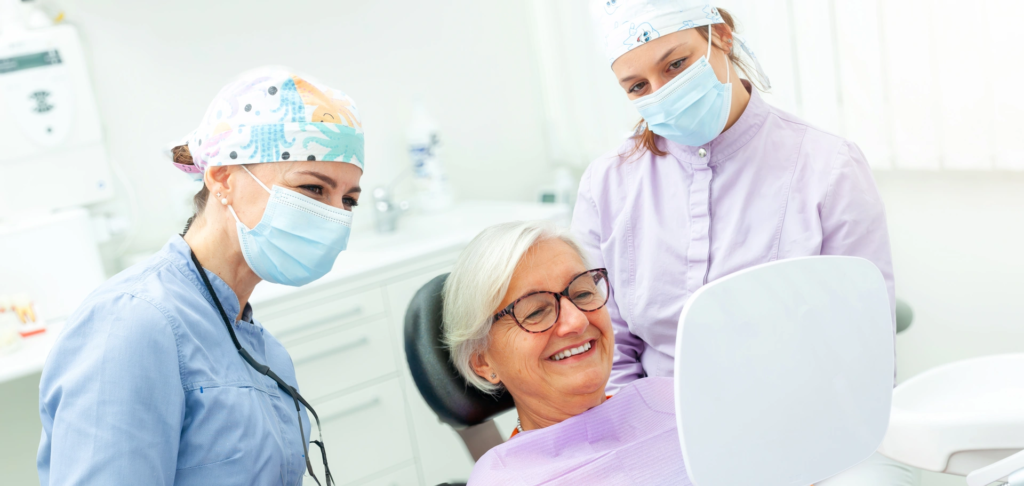 Starsza pacjentka uśmiechająca się do lustra podczas wizyty kontrolnej po wszczepieniu implantu zęba